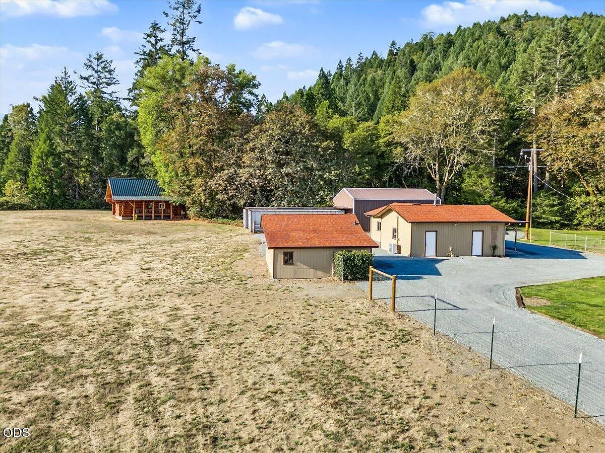 Aerial view across the finished outbuildings, log cabin, open ground, and wooded hillside